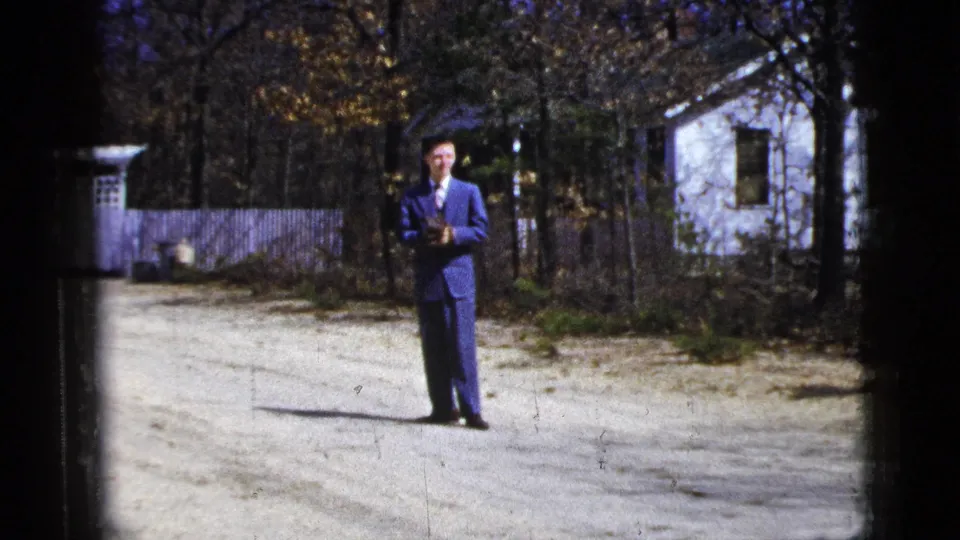 1956 8mm frame showing a man in a blue suit closer in view, standing by a gravel drive, fence, white house, and trees.
