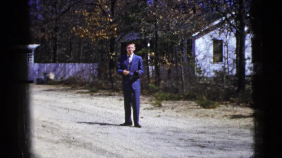 1956 New York 8mm frame with a man in a blue suit centered on a driveway, with a white house and autumn trees behind him.