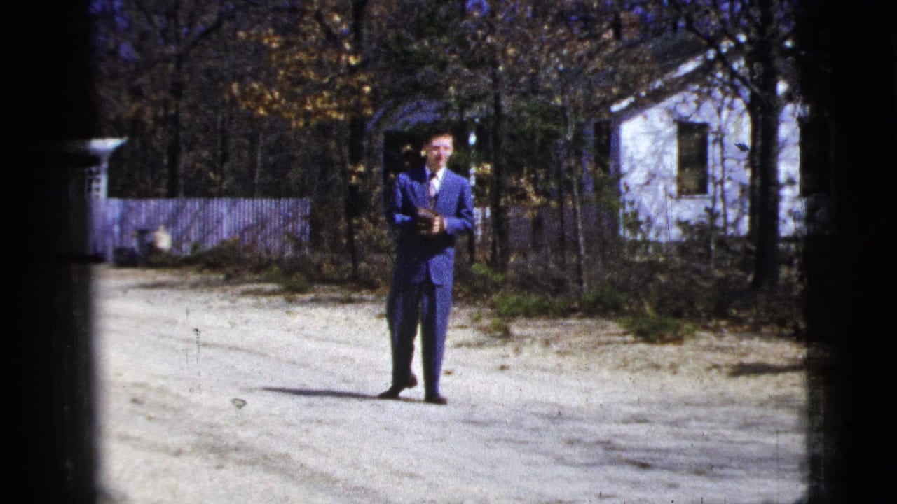 In 1956 New York City home-movie footage, a man in a blue suit stands on a dirt road with trees and houses behind him.