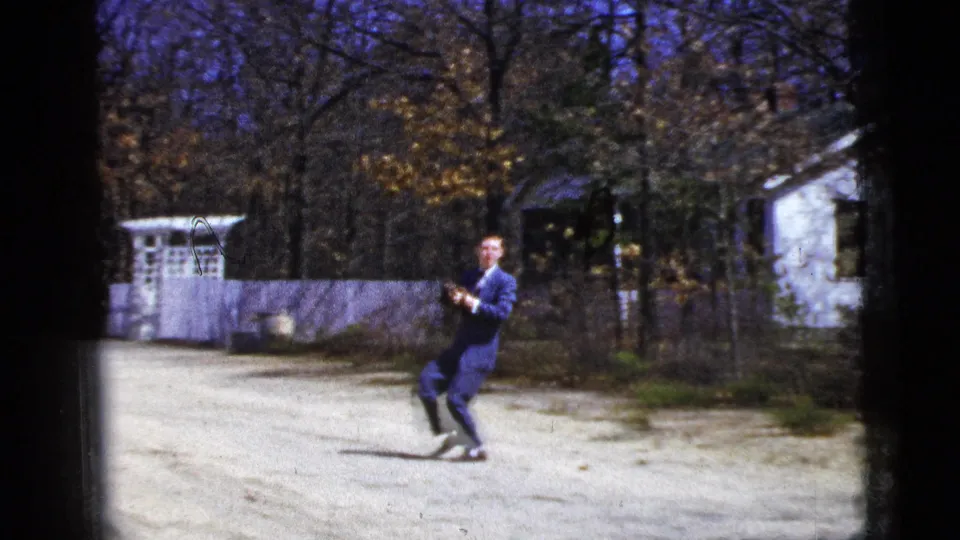 In the 1956 home-movie scene, the suited man leans back on the dirt road while tracking the ball with his glove raised.