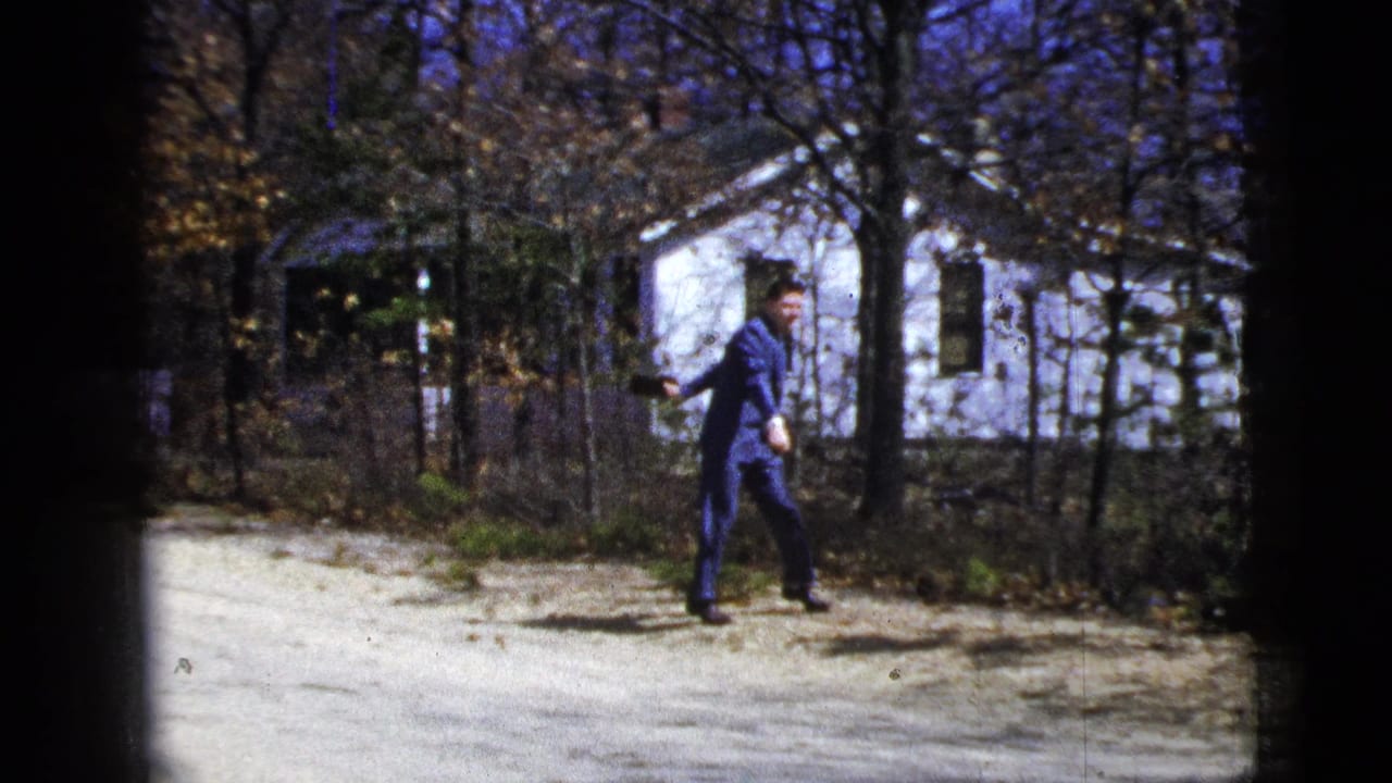 In 1956 New York City home-movie footage, a man in a blue suit moves along a dirt path in front of a white house and bare trees.