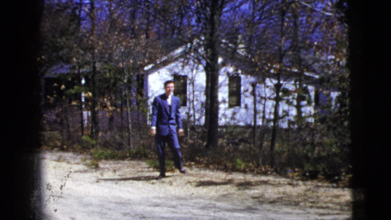 The suited man faces the camera on a dirt path, with a white residential house and leafless trees behind him.