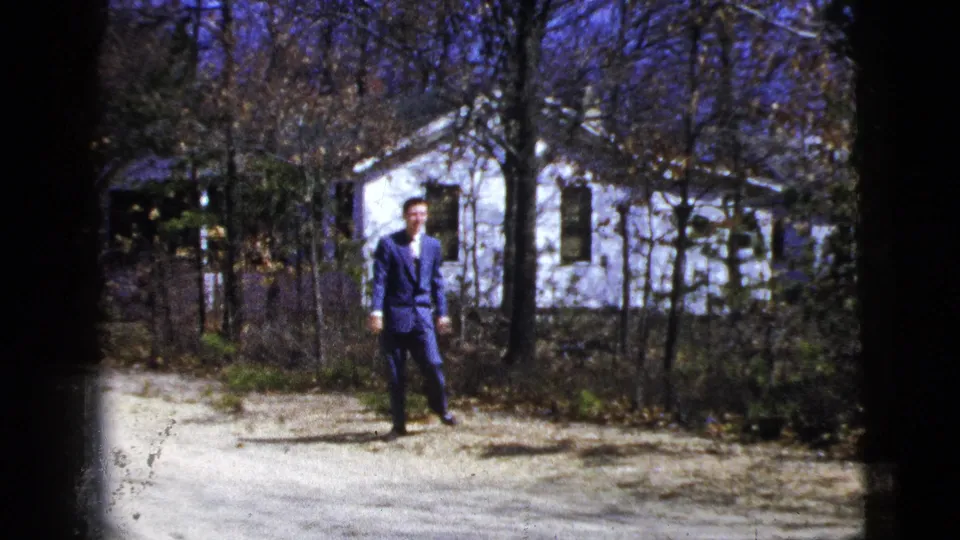 The suited man faces the camera on a dirt path, with a white residential house and leafless trees behind him.