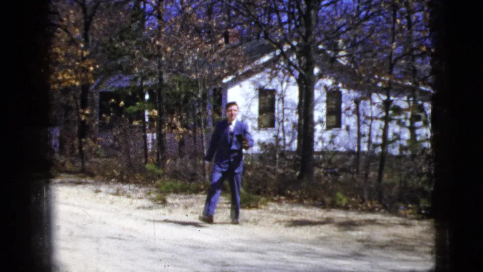 The man runs toward the camera in the residential outdoor scene, framed by bare branches, a fence, and the white house.