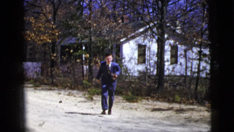 The suited man approaches the camera on the dirt path, smiling or animated, with the white house and autumn trees in the background.