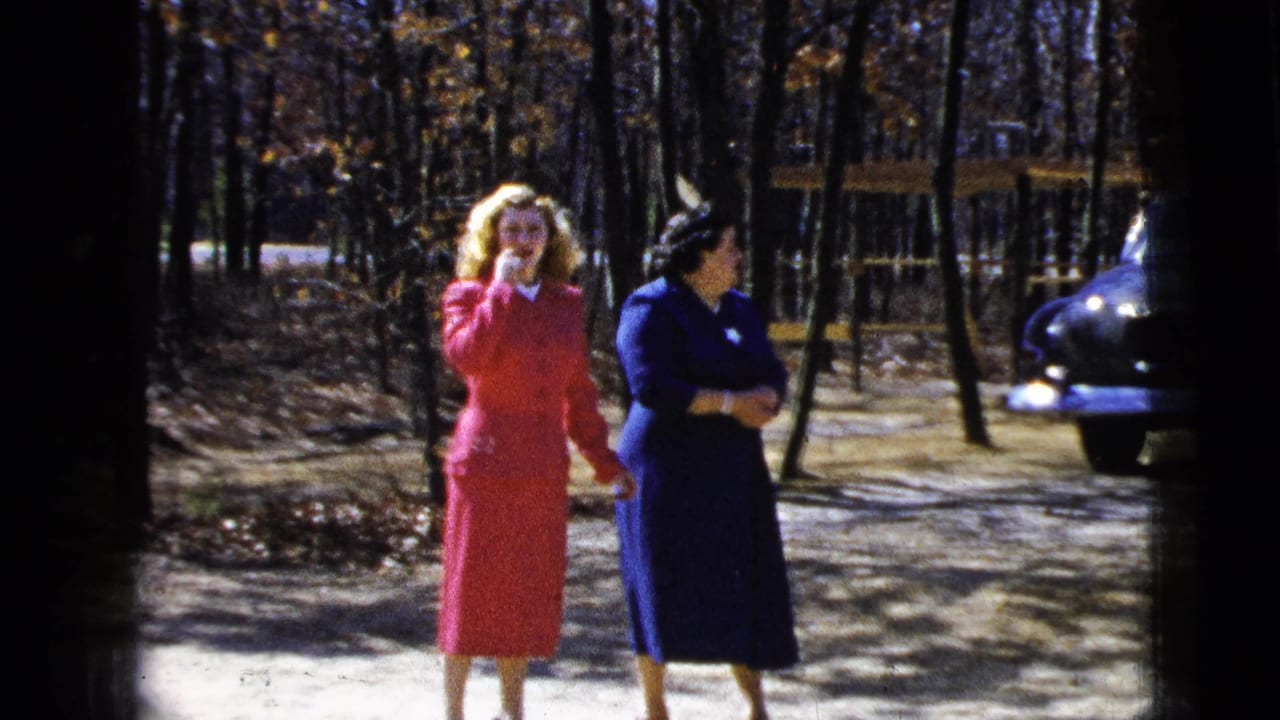 Two women in mid-1950s attire walk and talk outdoors in a wooded New York City area setting, with a parked car nearby.