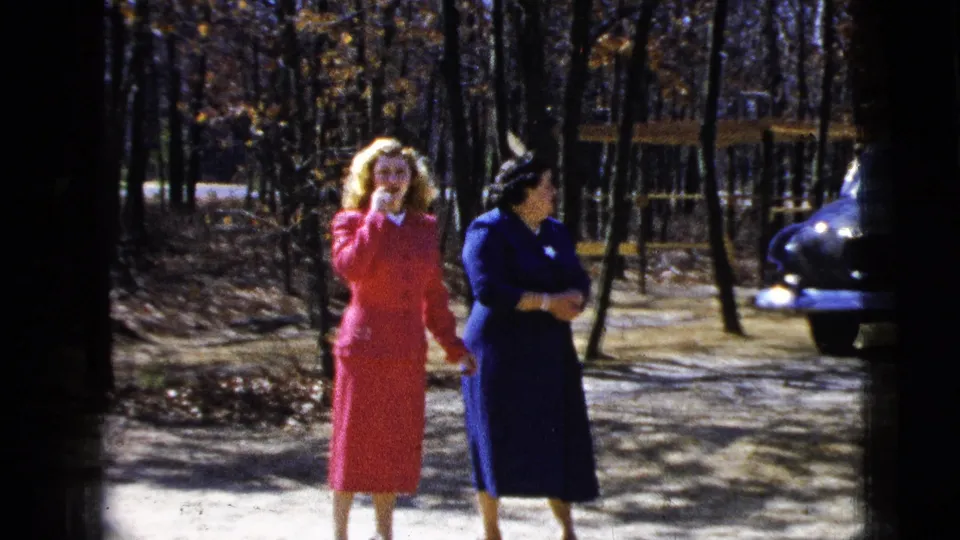 Two women in mid-1950s attire walk and talk outdoors in a wooded New York City area setting, with a parked car nearby.