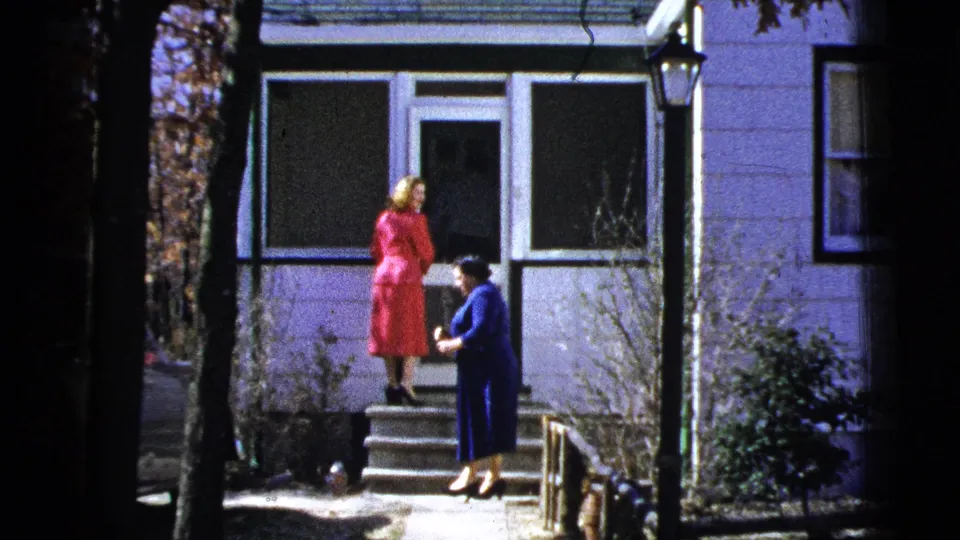 Two women in 1950s clothing pause near porch steps outside a light-colored house in the New York City area.