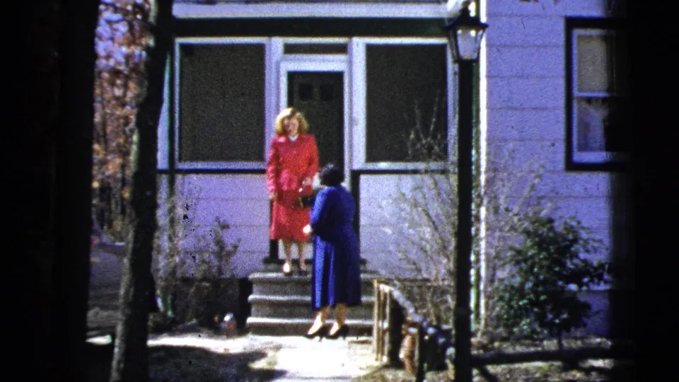 Outside a light-colored house, a woman in red stands on the steps while a woman in navy bends near the path.