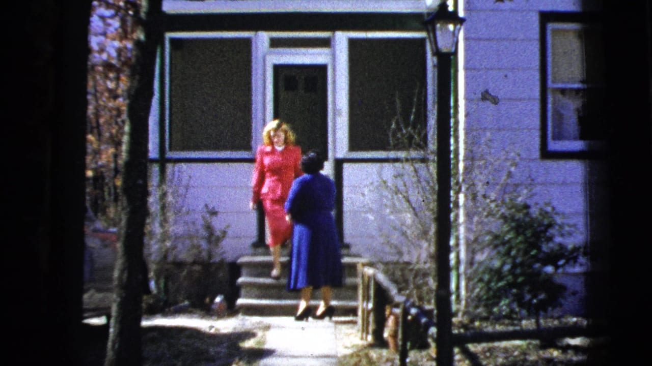 In a 1956 New York home-movie frame, a woman in a red suit faces another woman in blue at a residential doorway.