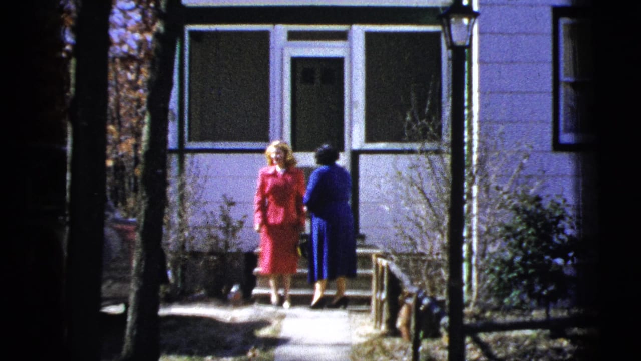 The two women remain outside the home, one in red and one in blue, framed by trees and the house front.