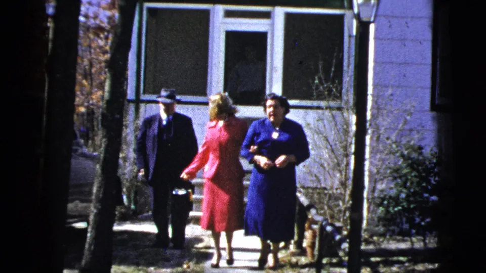 A suited man in a fedora walks beside two dressed-up women on a sunlit residential path in the 1956 home movie.