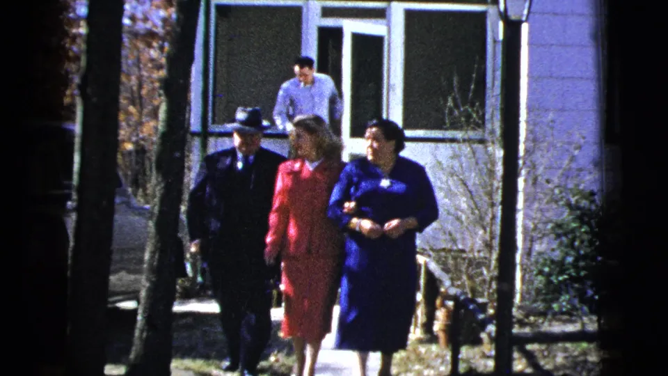 1956 New York home-movie frame showing three formally dressed adults walking outdoors from a light-colored house.