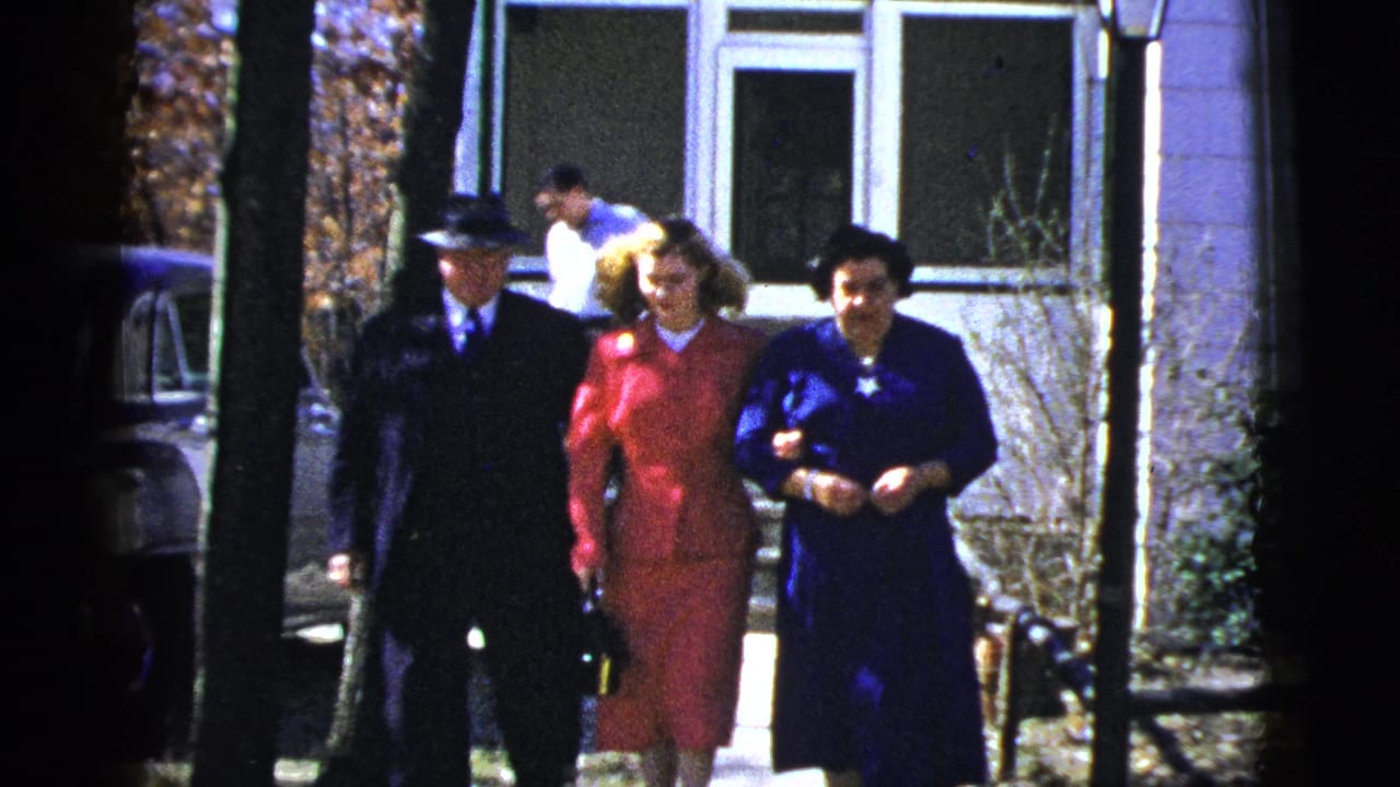 1956 New York frame of formally dressed adults moving along a residential walkway outside a house.