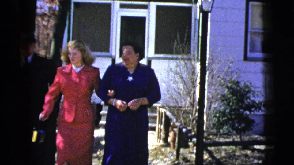 1956 New York home-movie still with two women in formal clothing walking close to a residential exterior.
