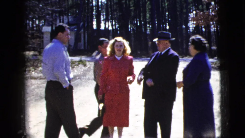 1956 New York frame showing adults in formal dress and a younger man in a light shirt talking outside a residence.