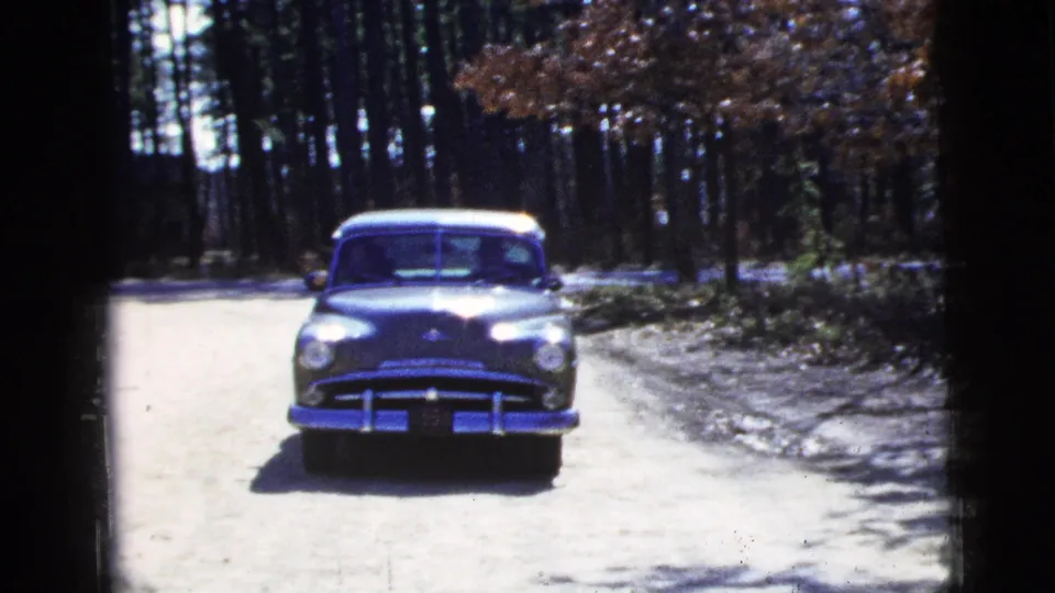 The dark mid-century sedan moves nearer on the wooded road, recorded with visible 8mm grain, border shadows, and bright exposure.