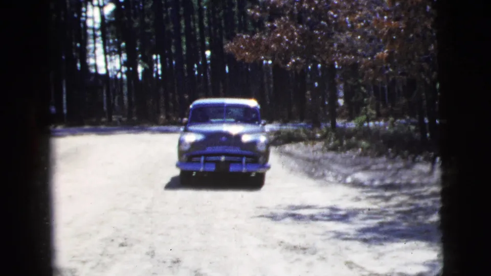 A 1950s sedan appears smaller on the sunlit road, with trees, brush, shadows, and 8mm film edges visible around the frame.
