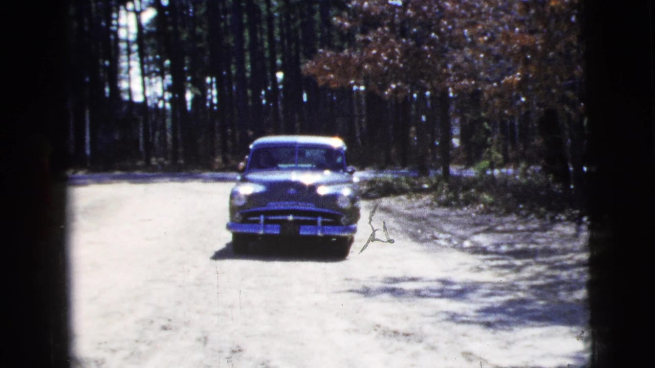 1956 8mm frame showing a dark green mid-century sedan approaching on a wooded dirt road in New York City metadata context.