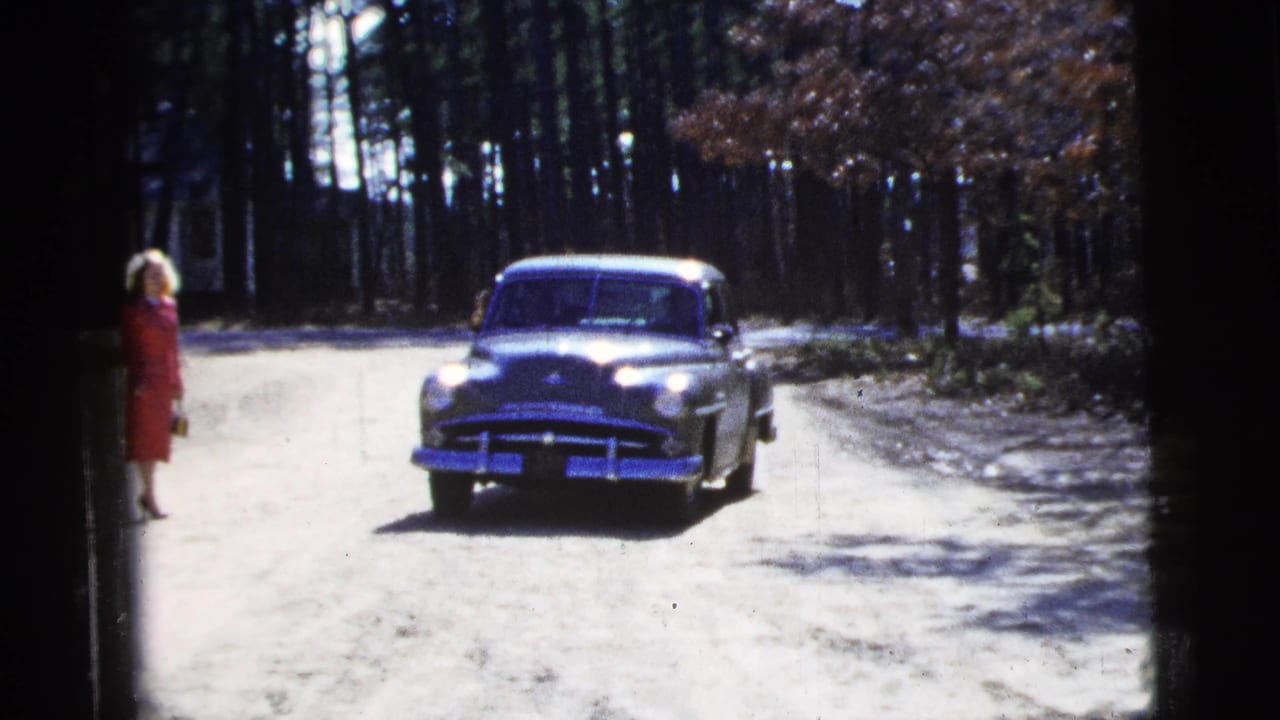 Woman in a red dress stands by the road as the green sedan approaches.