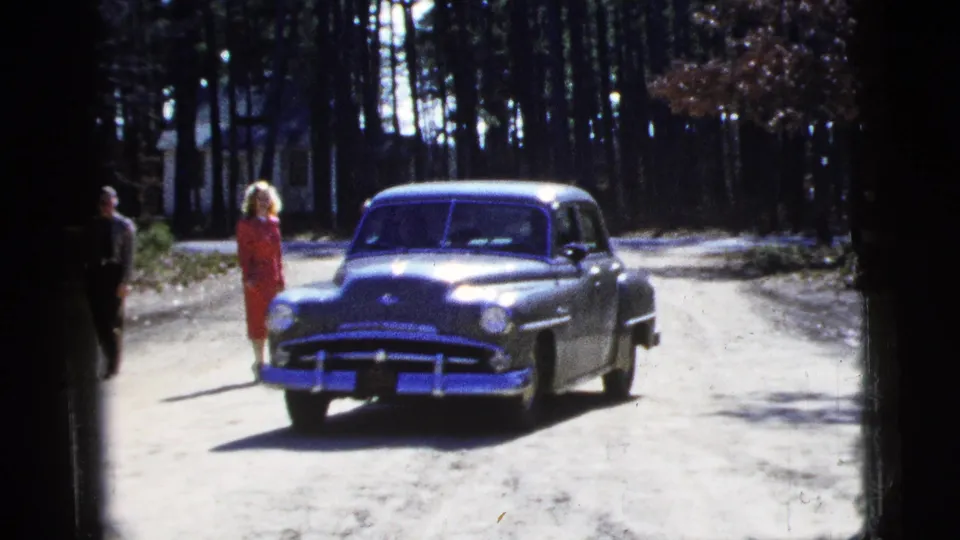 1956 8mm frame showing the green sedan moving past a woman in red, with another adult partly visible near the left edge.