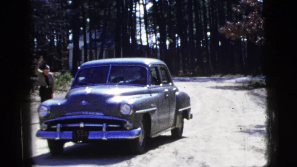 1956 8mm frame of the green sedan near the camera on a wooded dirt road, with an adult figure standing beside the road.