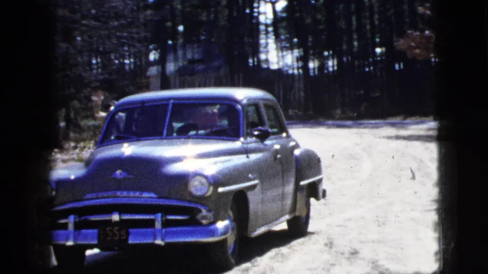 1956 New York home-movie frame of a silver sedan driving slowly toward camera on a wooded dirt road.