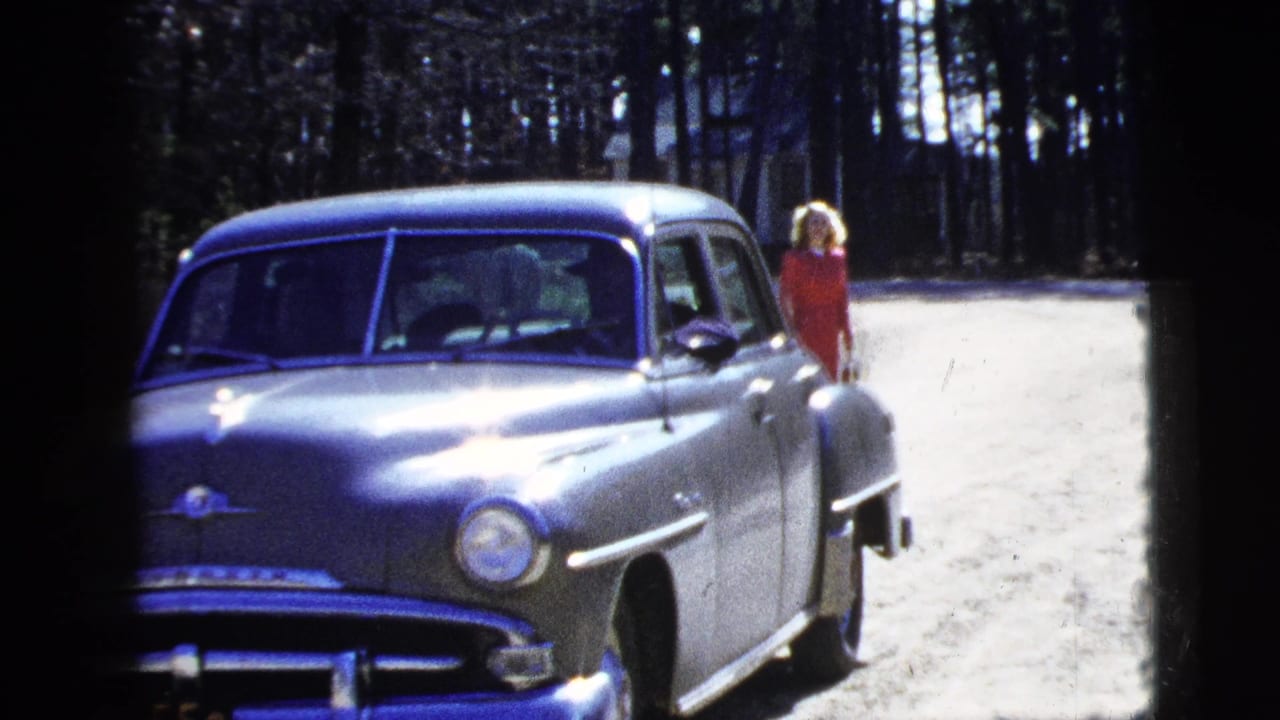 1956 New York frame showing a silver sedan on a dirt road with a woman in a red dress visible near the rear of the car.