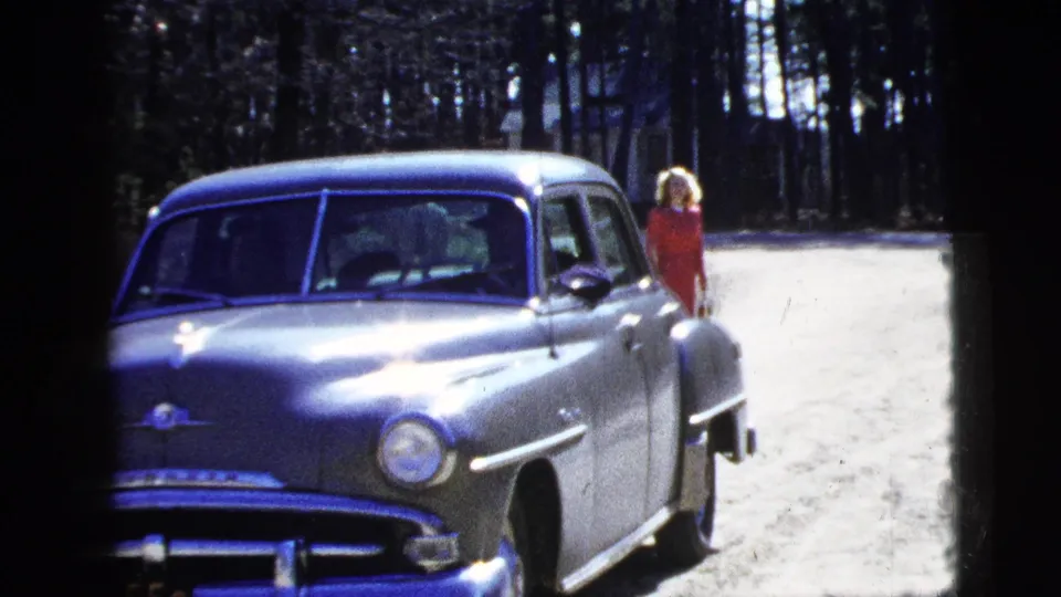 1956 New York frame showing a silver sedan on a dirt road with a woman in a red dress visible near the rear of the car.
