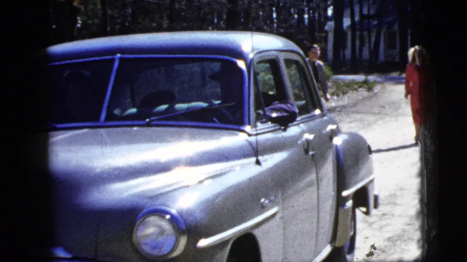 1956 New York home-movie frame with the silver sedan close to camera and two adults visible on the wooded roadside.