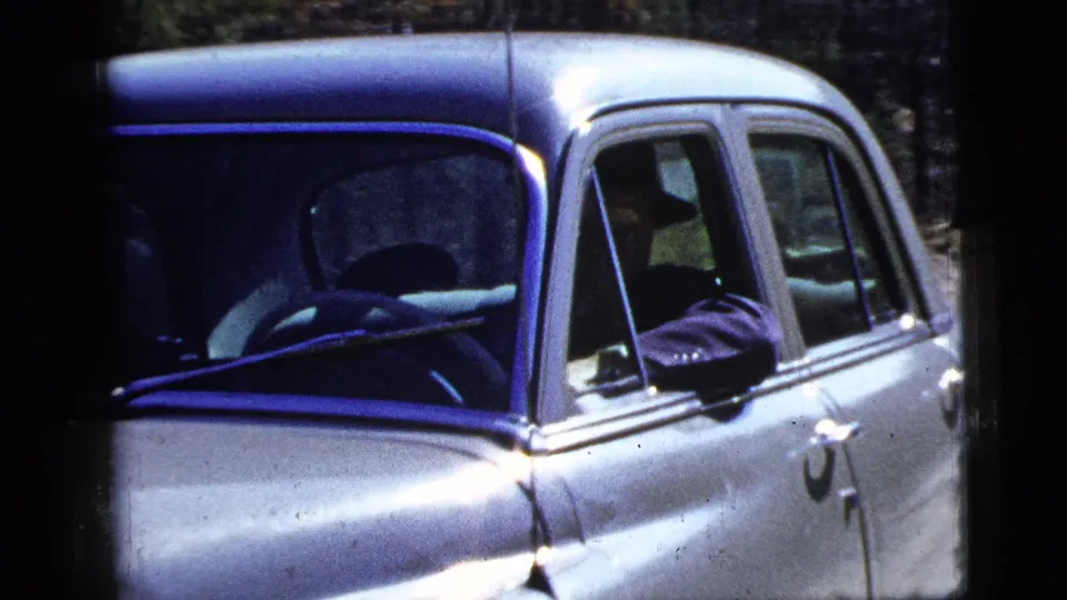 Close 1956 New York home-movie view of the silver sedan passing camera, with windshield, side window, and chrome trim visible.