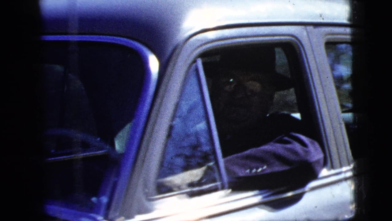 1950s 8mm frame showing a person seated in a light-colored sedan, viewed close through the side window in bright outdoor light.