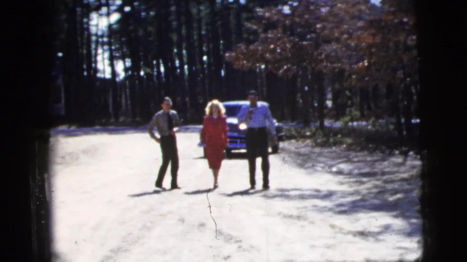 Three adults, including a woman in a red dress, walk toward the camera on a wooded road in New York City metadata, with a sedan behind them.