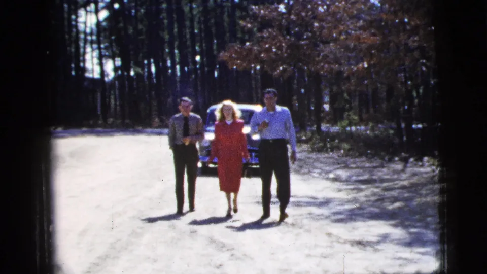 A woman in a red dress walks between two men as they near the camera in soft-focus 1956 New York City home-movie footage.