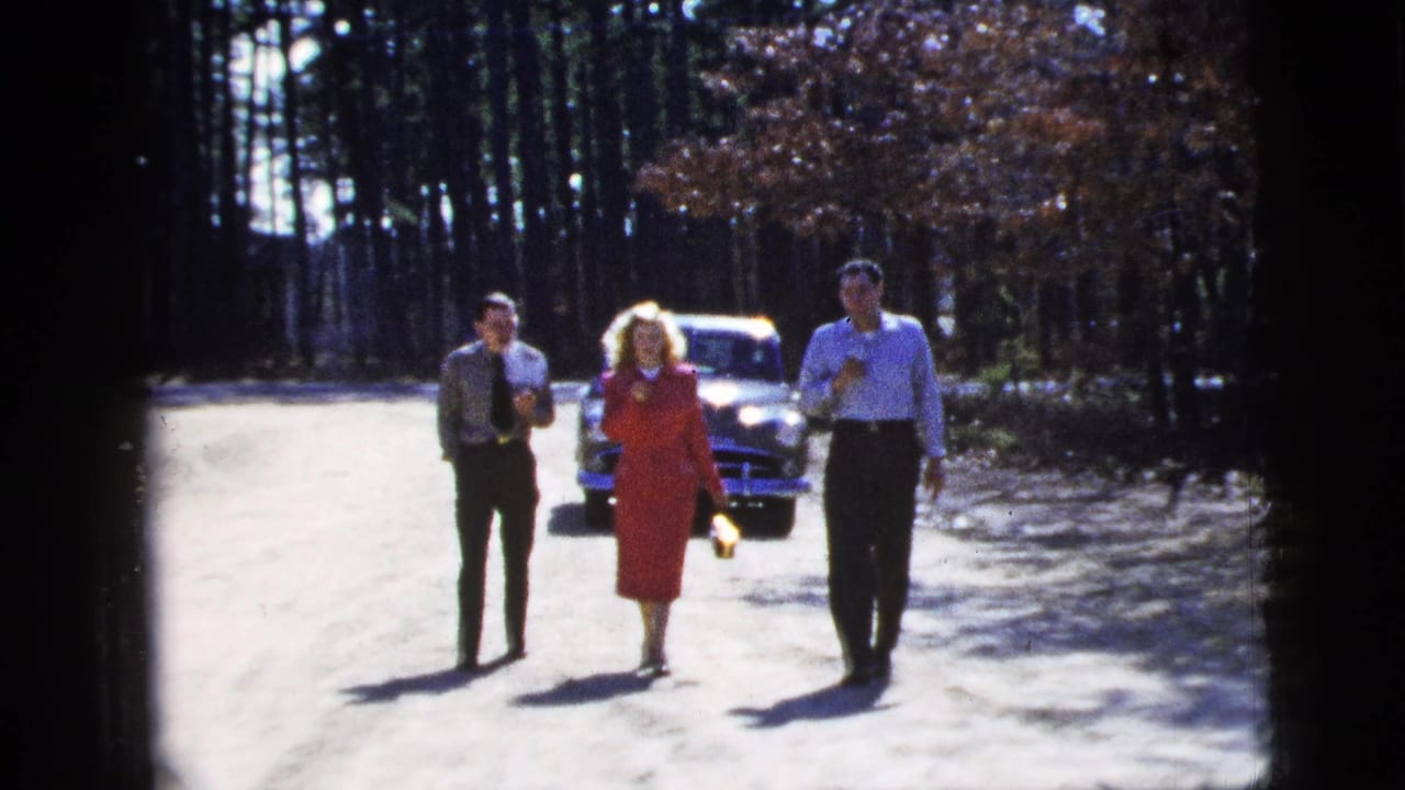 Three adults walk on a sunlit dirt road with a parked car and tall trees behind them.