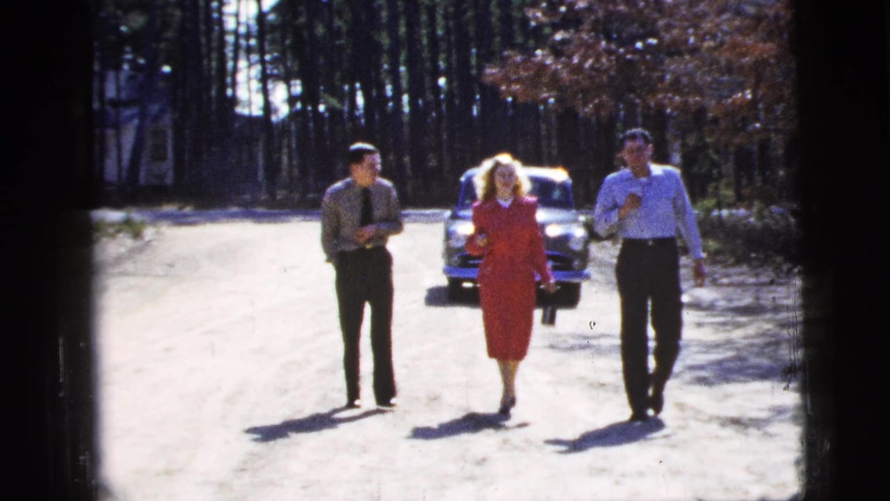 The group advances down the sunlit road in this 1956 New York City home-movie frame, with the woman in red centered between two men.
