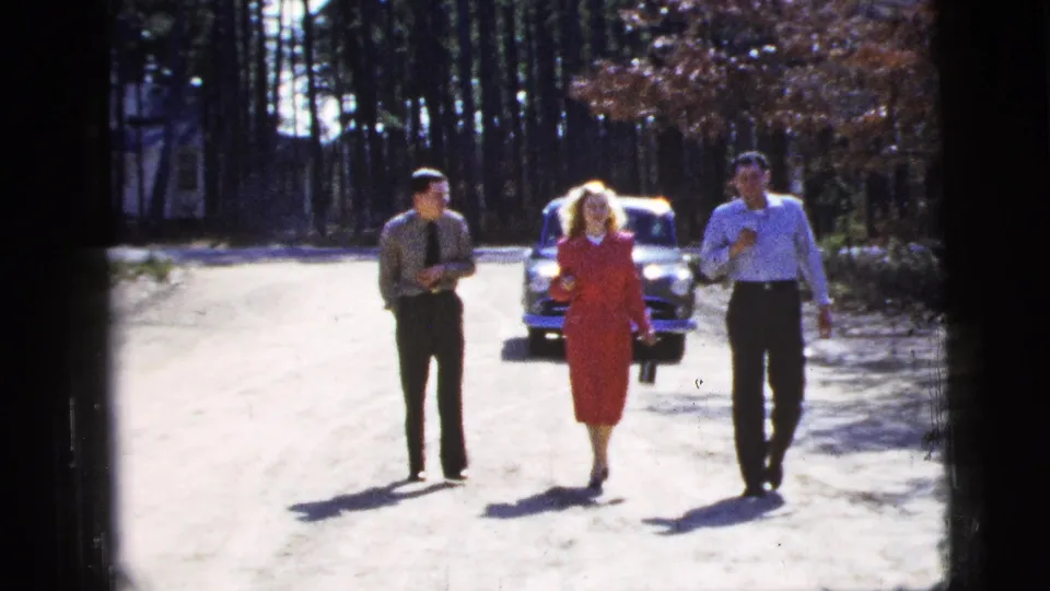 The group advances down the sunlit road in this 1956 New York City home-movie frame, with the woman in red centered between two men.
