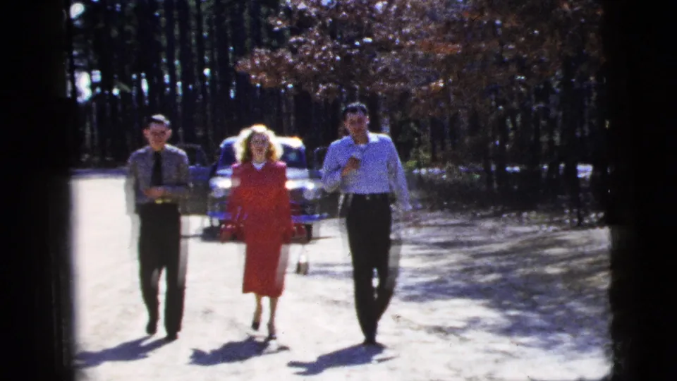 1956 New York City home-movie frame with three adults walking closer to the camera, the centered woman in red holding a small item and a car behind her.