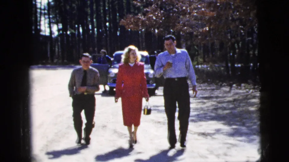 Near the end of the 1956 New York City clip, three adults walk close to camera, carrying drinks as a mid-century car sits behind them on the road.