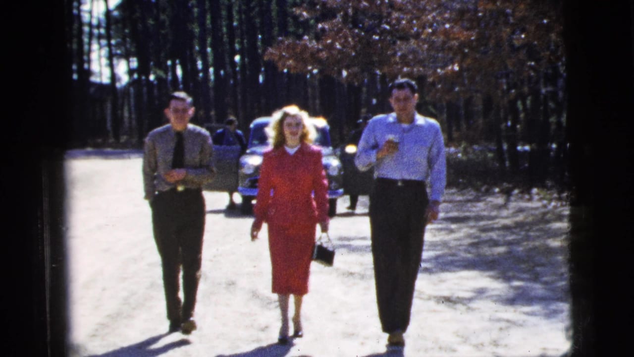 In this 1956 New York City home-movie frame, three adults walk toward the camera on a bright dirt path with trees and a classic car behind them.
