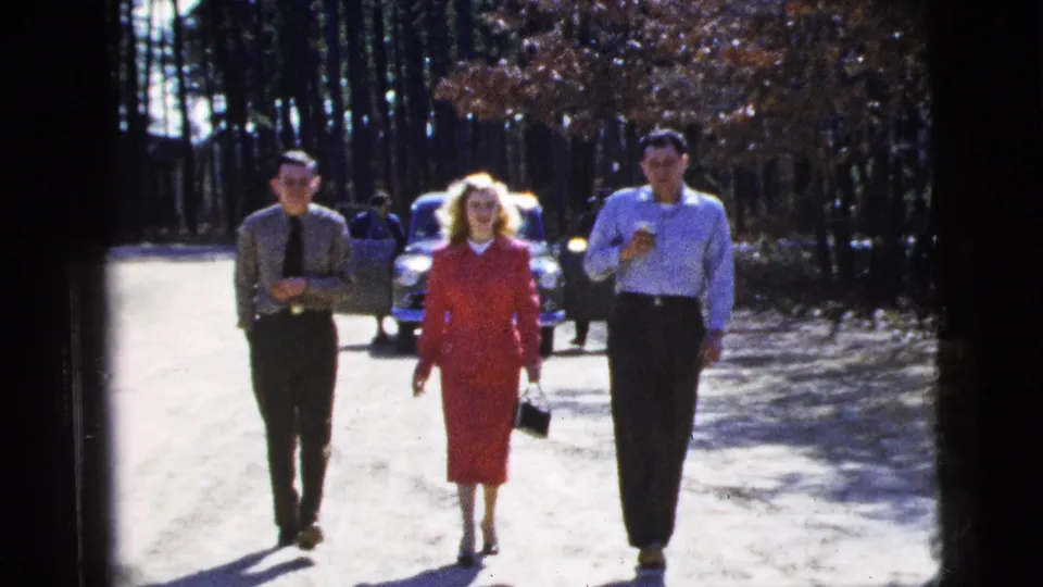 In this 1956 New York City home-movie frame, three adults walk toward the camera on a bright dirt path with trees and a classic car behind them.