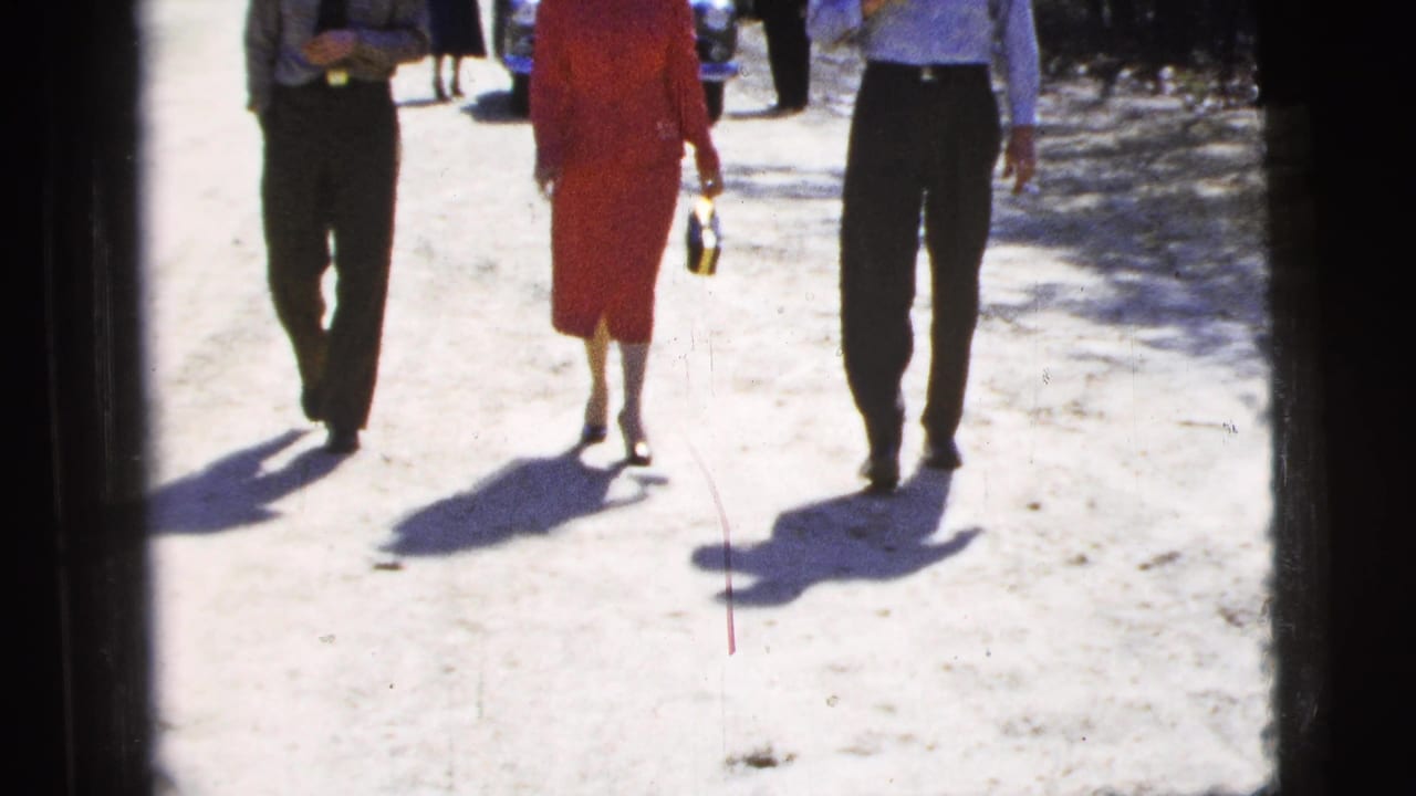 The camera frames the lower half of three adults walking along a sunlit dirt path, emphasizing their steps and shadows.