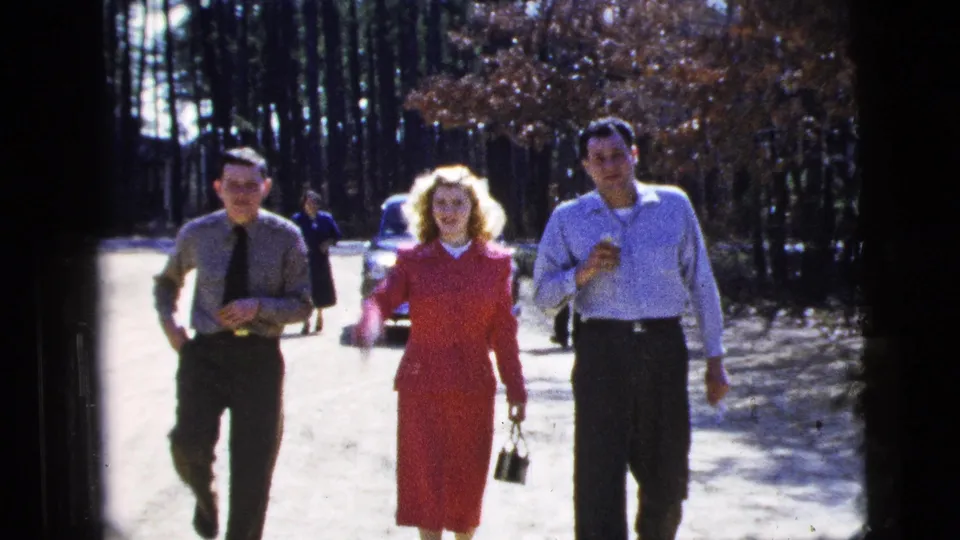 The group is closer to the camera, with the woman in a red dress between two men and parked cars visible along the wooded path.