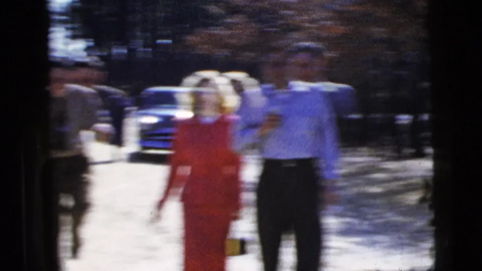 A motion-blurred 8mm frame shows the woman in red and a man walking past the camera near parked cars and trees.