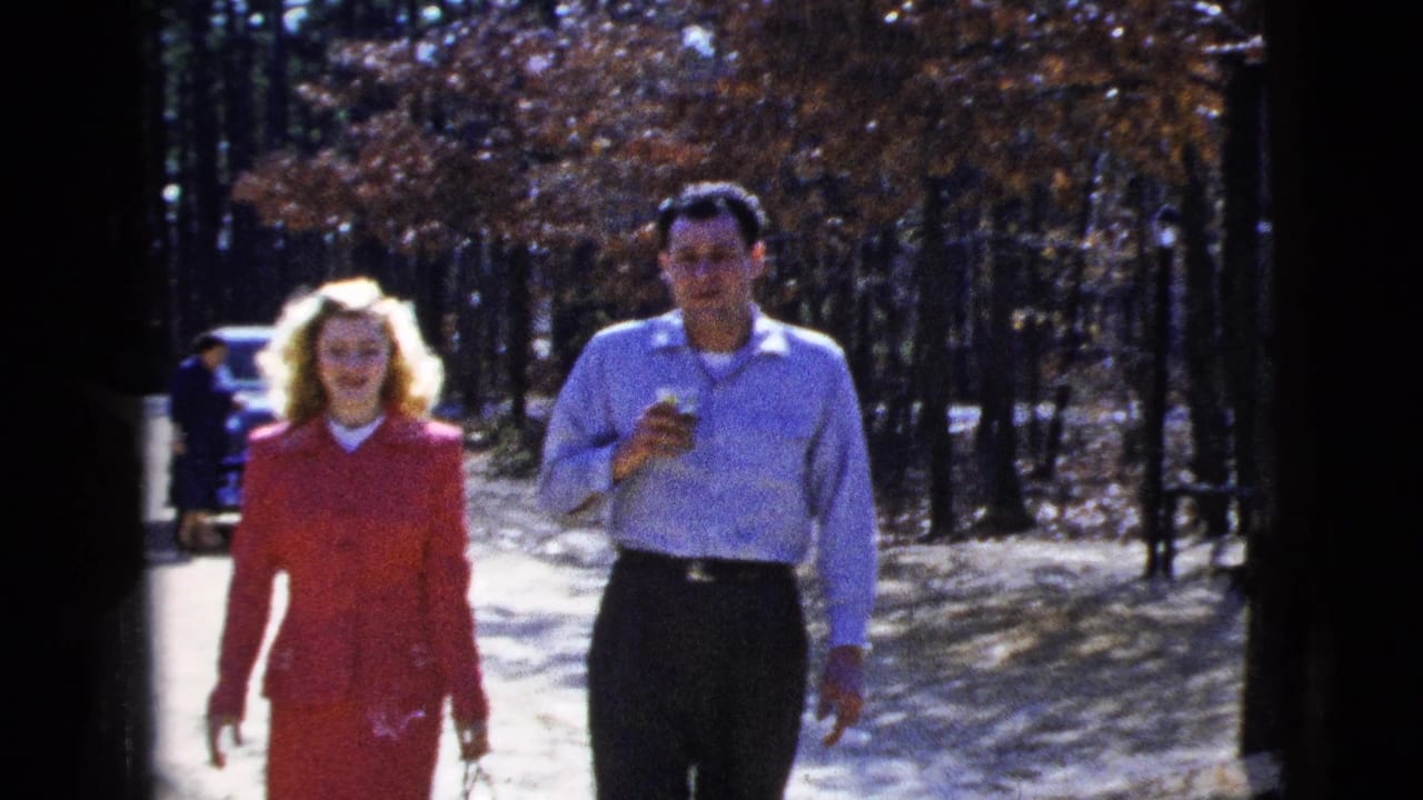 In 1956 New York City home-movie footage, a woman in a red suit and a man in a light shirt walk along a sunlit dirt road with trees behind them.