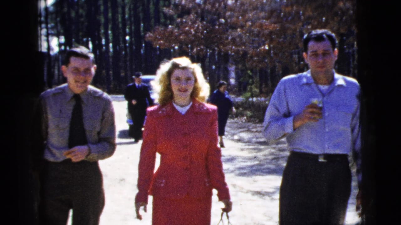 A 1956 New York City home-movie frame shows two adult men and a woman in a red suit walking toward the camera on a bright wooded road.