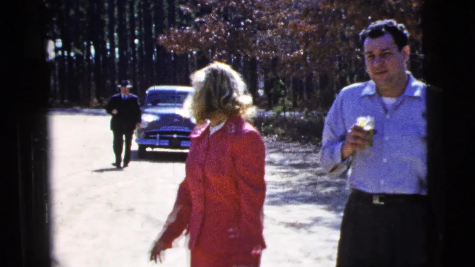 The woman in a red suit turns near the camera while a man holding a drink stands beside her, with a mid-century car and pedestrian visible on the road.