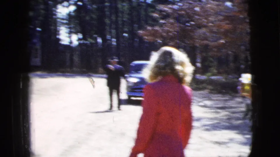 A woman in a red suit is seen from behind walking away on a wooded dirt road, with a car and distant figure visible in the 1956 New York City footage.