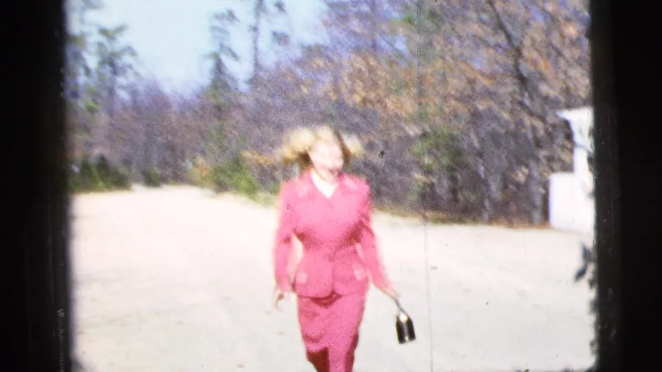 In supported 1956 New York City home-movie footage, a blonde woman in a pink suit approaches on a sunlit road.