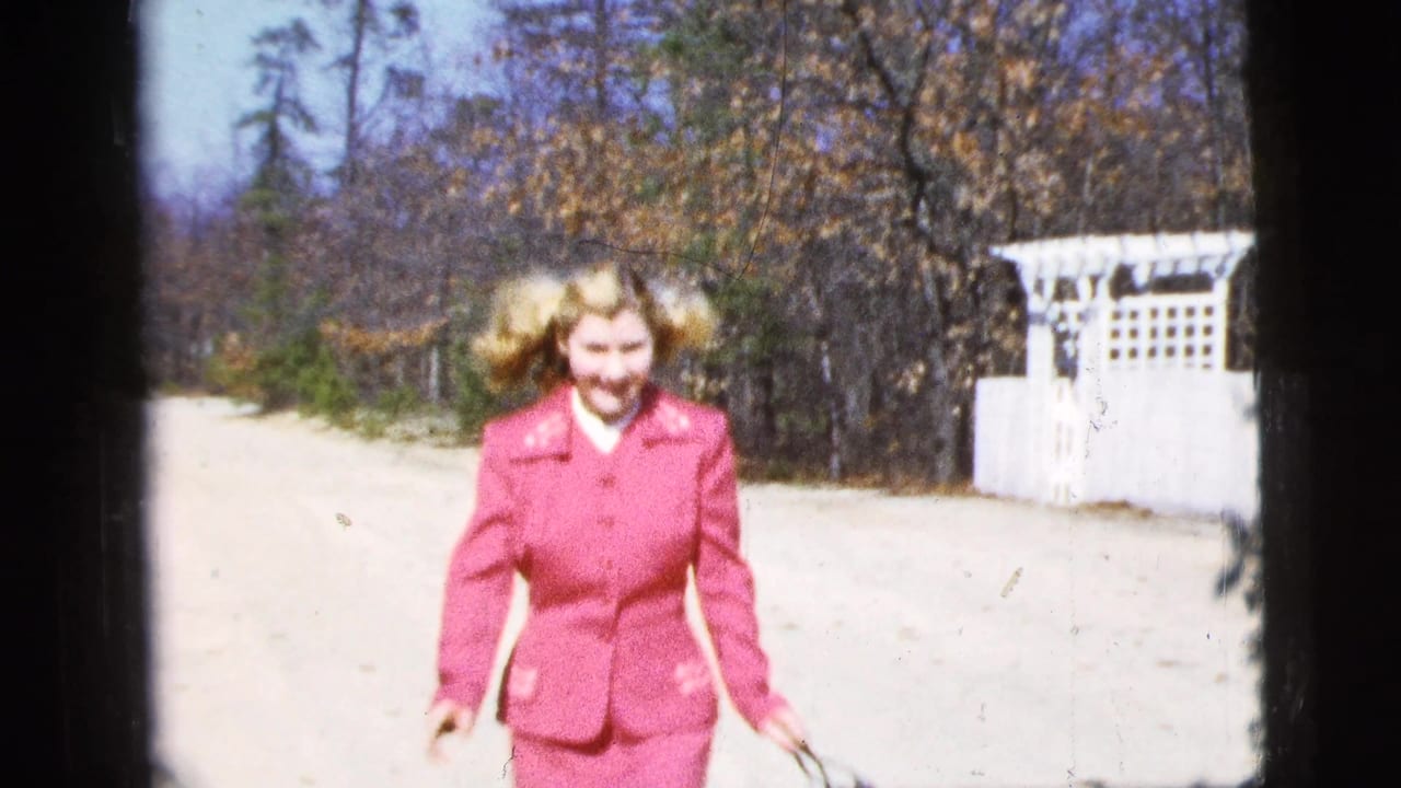 The woman smiles while skipping closer to the camera, with trees and a white fence visible behind her.